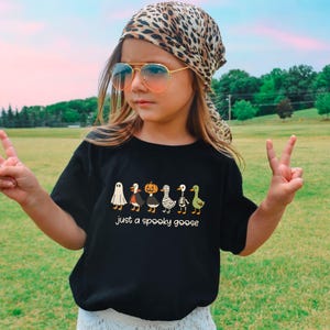 May include: A black t-shirt with a Halloween-themed graphic featuring cartoon geese dressed as a ghost, a vampire, a pumpkin, a skeleton, and a witch. The text "just a spooky goose" is below the geese. The person is wearing a leopard print scarf and sunglasses.
