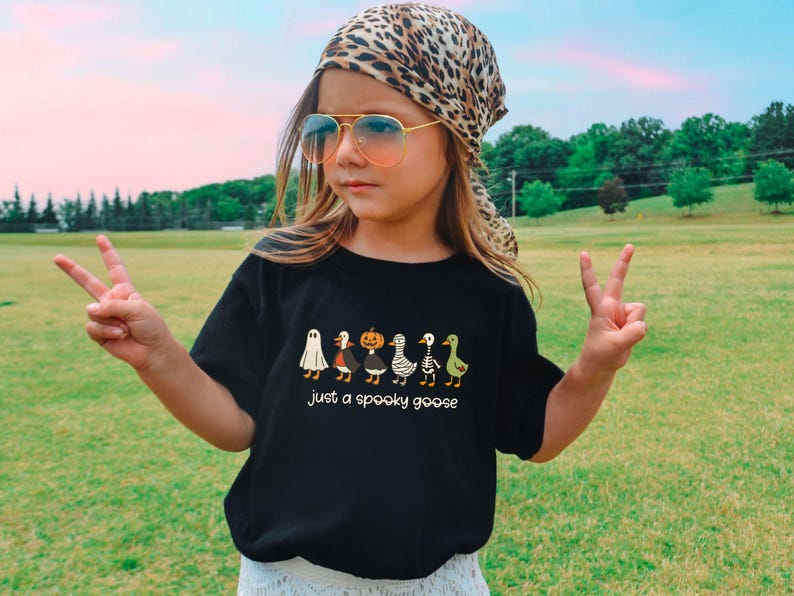 May include: A black t-shirt with a Halloween-themed graphic featuring cartoon geese dressed as a ghost, a vampire, a pumpkin, a skeleton, and a witch. The text "just a spooky goose" is below the geese. The person is wearing a leopard print scarf and sunglasses.