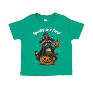 Peut inclure: T-shirt vert pour enfant avec un motif d'Halloween. Il repr&eacute;sente un raton laveur portant un chapeau et une cape de sorci&egrave;re, assis sur une citrouille sculpt&eacute;e et tenant une lanterne. Le texte "Spooky and Feral" est au-dessus du raton laveur.