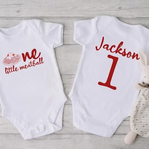 May include: Two white baby onesies. One has the text "one little meatball" in red, with a graphic of spaghetti and meatballs. The other onesie has the name "Jackson" and the number "1" in red. A stuffed bunny is next to the onesie.