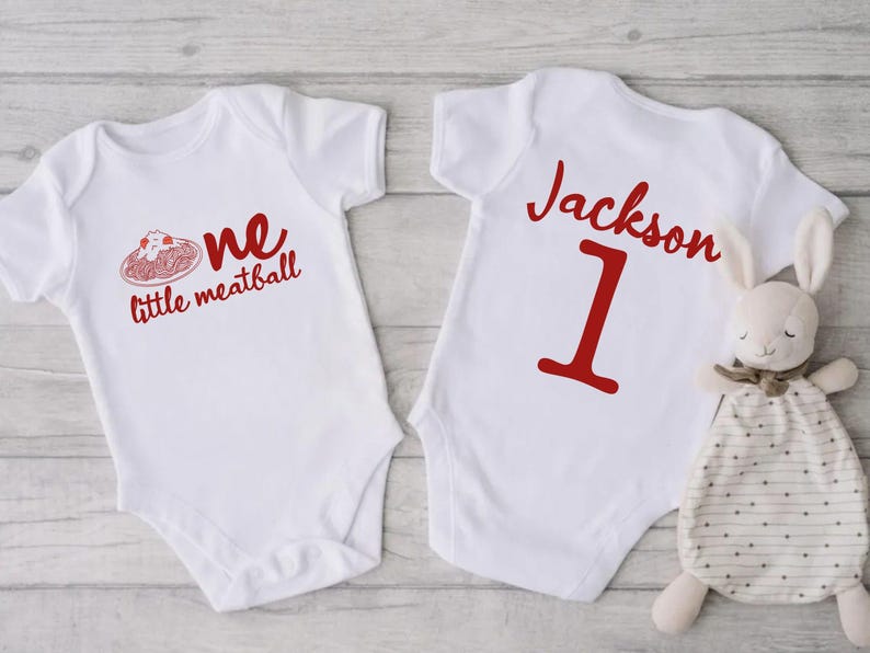 May include: Two white baby onesies. One has the text "one little meatball" in red, with a graphic of spaghetti and meatballs. The other onesie has the name "Jackson" and the number "1" in red. A stuffed bunny is next to the onesie.