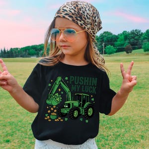May include: Black t-shirt with a green tractor graphic and the text "Pushin' My Luck". The design includes a tractor with a bucket full of shamrocks and a pot of gold, perfect for St. Patrick's Day.