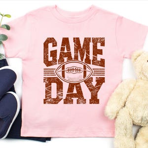 K&ouml;nnte beinhalten: Hellrosa T-Shirt mit einem verwitterten braunen Aufdruck "GAME DAY" mit einem Football in der Mitte. Das Shirt wird mit dunkelblauen Jeans, marineblauen Slip-On-Schuhen und einem Teddyb&auml;r pr&auml;sentiert.