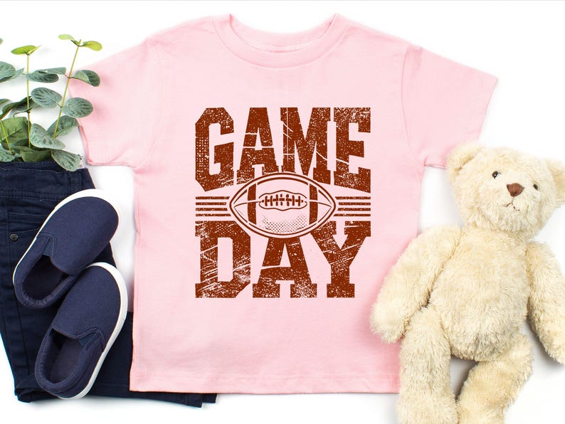 K&ouml;nnte beinhalten: Hellrosa T-Shirt mit einem verwitterten braunen Aufdruck "GAME DAY" mit einem Football in der Mitte. Das Shirt wird mit dunkelblauen Jeans, marineblauen Slip-On-Schuhen und einem Teddyb&auml;r pr&auml;sentiert.