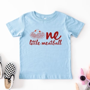May include: Light blue toddler t-shirt with the text "one little meatball" in red. The graphic features a plate of spaghetti and meatballs. A pair of black and white checkered slip-on shoes are next to the shirt.