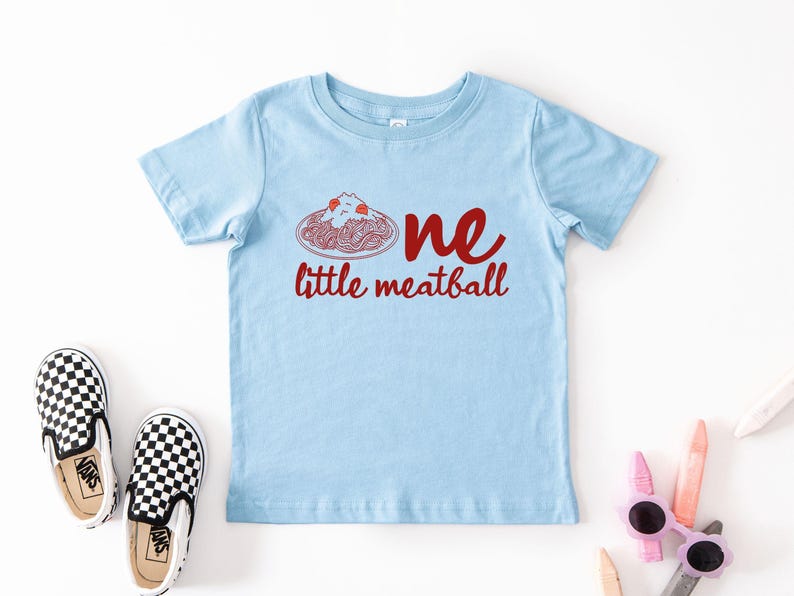 May include: Light blue toddler t-shirt with the text "one little meatball" in red. The graphic features a plate of spaghetti and meatballs. A pair of black and white checkered slip-on shoes are next to the shirt.