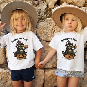 Peut inclure: Deux enfants portant des t-shirts blancs avec un motif d'Halloween. Les chemises pr&eacute;sentent un raton laveur avec un chapeau de sorci&egrave;re tenant une citrouille, avec le texte "Spooky and Feral". Ils se tiennent la main devant un mur de pierre.