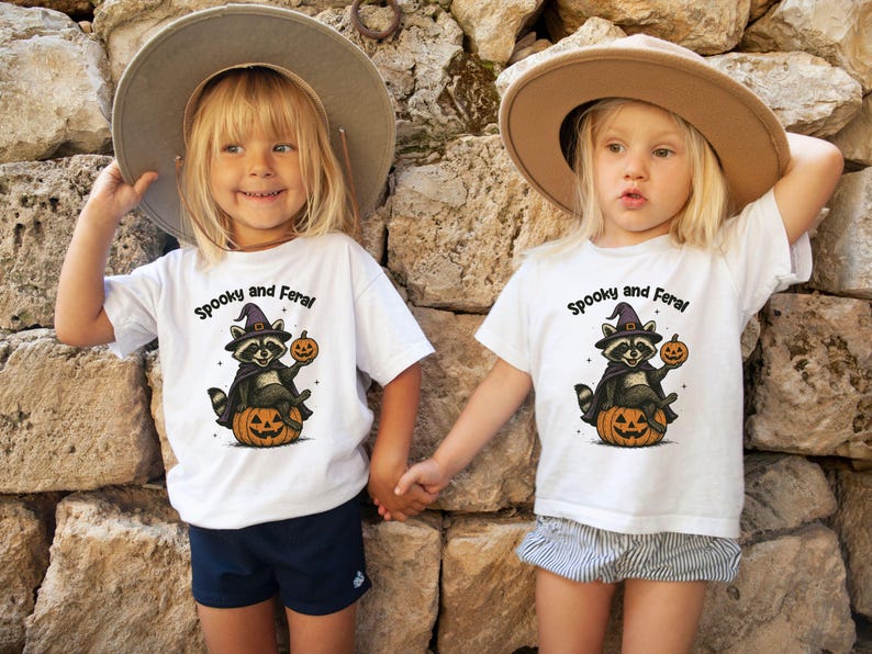 Peut inclure: Deux enfants portant des t-shirts blancs avec un motif d'Halloween. Les chemises pr&eacute;sentent un raton laveur avec un chapeau de sorci&egrave;re tenant une citrouille, avec le texte "Spooky and Feral". Ils se tiennent la main devant un mur de pierre.