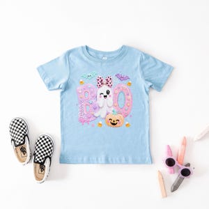 Puede incluir: Una camiseta de manga corta azul claro con un estampado gr&aacute;fico de la palabra BOO en letras rosas y azules, un fantasma gui&ntilde;ando un ojo con un lazo rosa de lunares, una calabaza naranja sonriente, murci&eacute;lagos peque&ntilde;os y caramelos.
