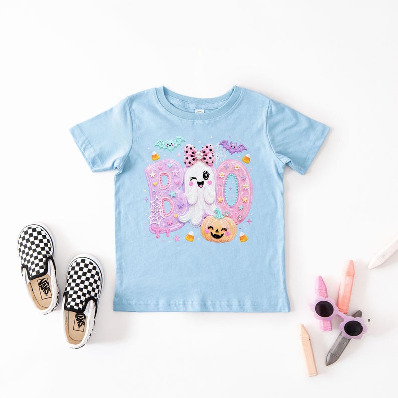Puede incluir: Una camiseta de manga corta azul claro con un estampado gr&aacute;fico de la palabra BOO en letras rosas y azules, un fantasma gui&ntilde;ando un ojo con un lazo rosa de lunares, una calabaza naranja sonriente, murci&eacute;lagos peque&ntilde;os y caramelos.