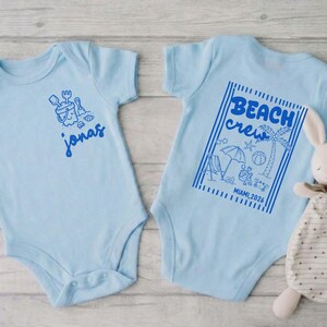 Op de afbeelding: Lichtblauwe babybody met een strandthema. E&eacute;n body heeft de naam "Jonas" in blauw. De andere heeft een "Beach Crew" ontwerp met een palmboom, parasol en de tekst "Miami 2024". Een knuffelkonijn ligt naast de body.