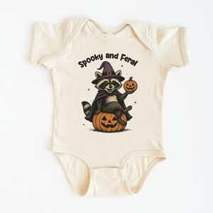 Peut inclure: Body b&eacute;b&eacute; cr&egrave;me avec un motif sur le th&egrave;me d'Halloween. L'illustration repr&eacute;sente un raton laveur portant un chapeau de sorci&egrave;re et une cape, assis sur une citrouille sculpt&eacute;e et tenant une petite citrouille. Le texte dit "Spooky and Feral."