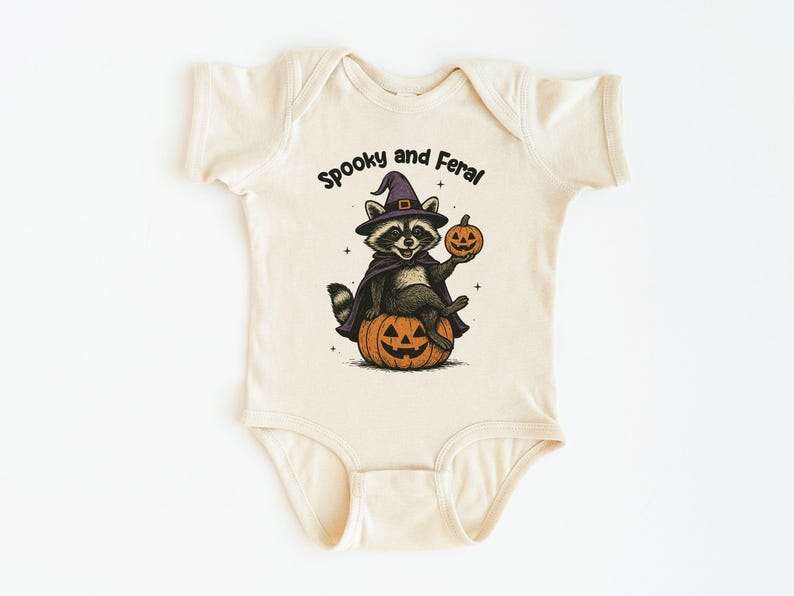 Peut inclure: Body b&eacute;b&eacute; cr&egrave;me avec un motif sur le th&egrave;me d'Halloween. L'illustration repr&eacute;sente un raton laveur portant un chapeau de sorci&egrave;re et une cape, assis sur une citrouille sculpt&eacute;e et tenant une petite citrouille. Le texte dit "Spooky and Feral."