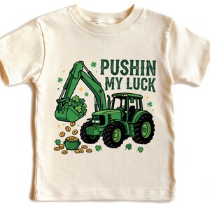 May include: Cream-colored toddler t-shirt featuring a green tractor with a bucket full of shamrocks and gold coins. The text "PUSHIN MY LUCK" is printed above the tractor, with shamrock and star accents.