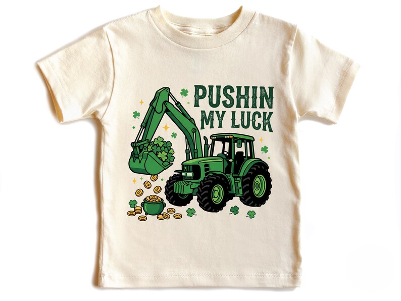 May include: Cream-colored toddler t-shirt featuring a green tractor with a bucket full of shamrocks and gold coins. The text "PUSHIN MY LUCK" is printed above the tractor, with shamrock and star accents.