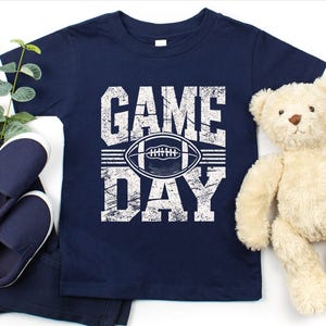 K&ouml;nnte beinhalten: Marineblaues T-Shirt mit den Worten "GAME DAY" in abgenutzten wei&szlig;en Buchstaben und einer Football-Grafik. Das Shirt wird mit marineblauen Schuhen, einer Hose und einem Teddyb&auml;r kombiniert.
