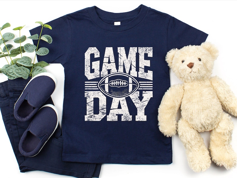 K&ouml;nnte beinhalten: Marineblaues T-Shirt mit den Worten "GAME DAY" in abgenutzten wei&szlig;en Buchstaben und einer Football-Grafik. Das Shirt wird mit marineblauen Schuhen, einer Hose und einem Teddyb&auml;r kombiniert.