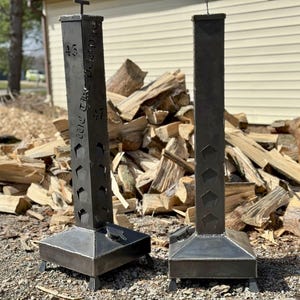 Może przedstawiać: Dwa ciemne metalowe kominy do palenisk zewnętrznych z kwadratową podstawą i wysokim, prostokątnym korpusem. Kominy mają wycięte wzory i tekst z boku. Stoją na żwirowej powierzchni, z tyłu stos drewna opałowego.