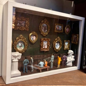 Può includere: Una scatola ombra con cornice bianca con figure in miniatura e foto incorniciate. Le foto sono in cornici dorate ornate. Lo sfondo è di colore verde scuro. Le figure in miniatura sono in piedi e sedute di fronte a un muro marrone scuro.