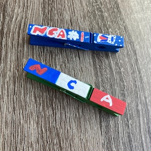 May include: Two wooden clothespins painted in red, white, and blue with the letters "NCA" and "CA" painted on them.