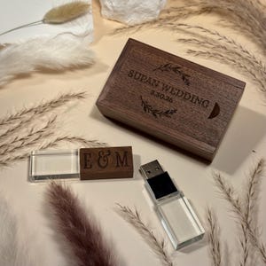 May include: A wooden USB drive and case set. The case is engraved with "Supan Wedding 3.30.25" and decorative elements. A second wooden piece is engraved with "E & M". The USB drive has a clear, rectangular body.