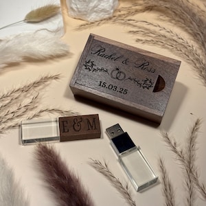 Puede incluir: Una caja de madera grabada con los nombres "Rachel & Ross" y la fecha "15.03.25". Una unidad USB con una carcasa de plástico transparente y una unidad USB de madera con las letras "E & M" también están presentes. Los artículos están sobre una superficie beige.
