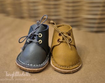 May include: A pair of miniature leather boots in contrasting colors. One is a muted blue-gray, the other a warm tan. Both have laces, stitched soles, and gold-colored eyelets. The boots are designed for dolls.