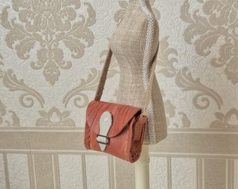 May include: A small, reddish-brown leather crossbody bag with a flap closure and a white accent. The bag has a textured design and a buckle closure. The strap is a matching brown color. The bag is displayed on a mannequin.