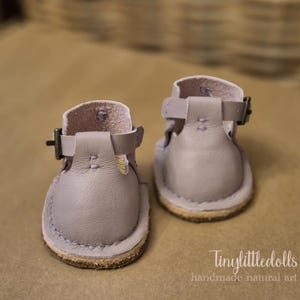 May include: A pair of lavender leather sandals with buckle closures. The small shoes have a rounded toe and a stitched sole. The sandals are designed for dolls and are handmade.