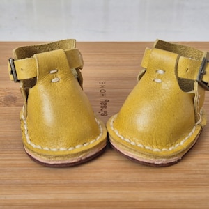 May include: A pair of small, yellow leather sandals with buckle closures. The sandals have white stitching around the edges and a pink sole. The sandals are sitting on a wooden surface.
