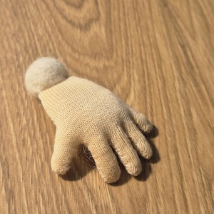 May include: A small, beige knitted glove with a rounded, fuzzy top. The glove has a hand shape with fingers and a wrist cuff. The item is lying on a wooden surface.