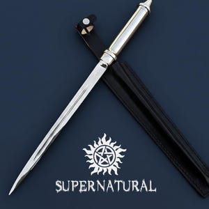 Könnte beinhalten: Ein silberfarbener Dolch mit spitzer Klinge und dekorativem Griff, der auf einer dunkelblauen Oberfläche liegt. Eine schwarze Lederscheide befindet sich hinter dem Dolch. Das Bild enthält den Text "SUPERNATURAL" und ein Sonnenstrahl-Pentagramm-Symbol.
