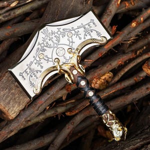 May include: A decorative hammer with a silver head featuring an intricate floral design and gold accents. The handle is wrapped in black leather and brown leather with gold detailing. The hammer rests on a bed of wooden branches.