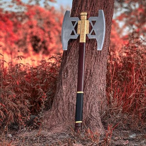 May include: A double-bladed axe with a silver head and a gold-colored central design. The handle is dark brown with a black grip and gold accents. The axe is leaning against a tree trunk in a forest setting.