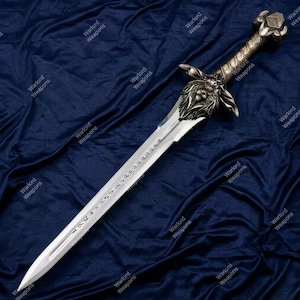 May include: A silver and bronze sword with a lion's head design on the hilt. The blade has etched text and a pointed tip. The sword is displayed on a dark blue velvet surface. The text "Warlord Weapons" is repeated around the image.