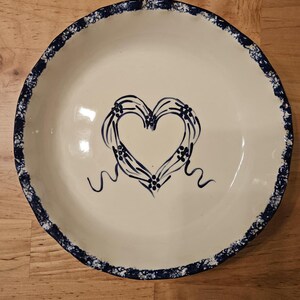May include: Cream-colored ceramic serving bowl with a blue heart design in the center. The heart is outlined with blue lines and adorned with small floral accents. The rim of the bowl has a blue speckled border.