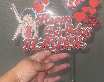 May include: A clear acrylic cake topper with a Betty Boop graphic, red hearts, and the words "Happy Birthday HAYLIE" in red. A heart-shaped element with the number 17 is also included. This topper is designed for a birthday celebration.