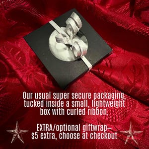 a small, lightweight box with a curled ribbon, placed on a red fabric background. The text indicates that the box is a gift, and it has been described as a "super secure packaging" with a "curled ribbon" inside. The image also includes a price tag of $5 extra, which can be chosen at checkout.