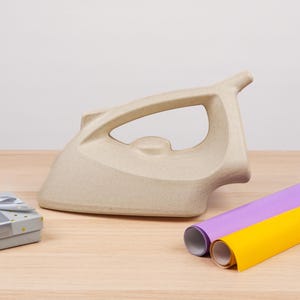 Puede incluir: Una plancha beige con un asa grande descansa sobre una superficie de madera. Junto a ella hay dos rollos de papel de colores, uno morado y otro amarillo. También hay una pequeña caja de regalo con un lazo plateado.