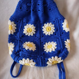May include: A blue crocheted tote bag with white and yellow daisy flower designs. The bag has two long straps.