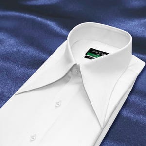 Puede incluir: Una camisa de vestir blanca y nítida con un cuello ancho de puntas y una tapeta de botones frontal. La etiqueta interior del cuello muestra el texto John Cloth London y Green. La camisa presenta un acabado liso, estructurado y botones.