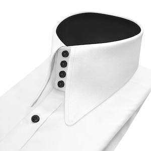 Può includere: Primo piano di una camicia bianca con colletto alto e arrotondato. Il colletto ha un interno nero ed è fissato con quattro bottoni neri. Un singolo bottone nero è visibile sulla paramontura della camicia.