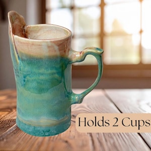 Puede incluir: Taza de cerámica hecha a mano con un diseño asimétrico único. La taza presenta un esmalte turquesa y marrón, con un asa curvada. El texto "Holds 2 Cups" es visible en la imagen. Ideal para café, té u otras bebidas.
