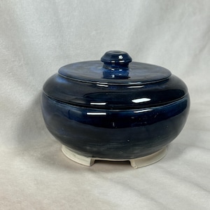 May include: A blue ceramic jar with a lid, featuring a dark blue glaze with white streaks. The jar has a round body and sits on three small feet.