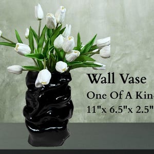 May include: A black, glossy wall vase with a unique, textured design, holding a bouquet of white tulips with green leaves. The vase measures 11" x 6.5" x 2.5". The text on the image reads "Wall Vase One Of A Kind".