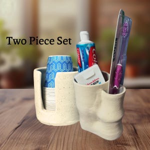 Peut inclure: Un ensemble de deux pièces en céramique blanc cassé moucheté pour la salle de bain. Une pièce est un porte-gobelet contenant une pile de gobelets en papier à motifs bleus et blancs. L'autre est un support compartimenté avec dentifrice et brosse.