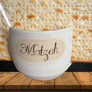 May include: A white ceramic bowl with the word "Matzah" written on it in black calligraphy. The bowl is sitting on a wooden surface in front of a piece of matzah.