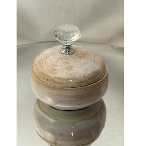 Könnte beinhalten: Eine kleine, runde Keramik-Schmuckdose mit einem gesprenkelten Überzug in Beige- und Weißtönen. Der Deckel hat einen durchsichtigen Kristallknopf.