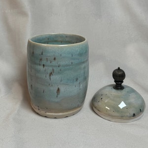 Könnte beinhalten: Eine Keramikdose mit Deckel. Die Dose ist hellblau mit braunen Flecken und einem weißen Boden. Der Deckel ist hellblau mit braunen Flecken und hat einen Metallknopf.