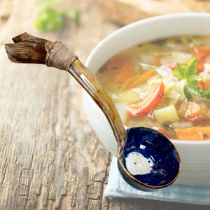 May include: A handcrafted ceramic soup ladle with a dark blue bowl and a wooden handle. The handle is wrapped with twine. The ladle is next to a bowl of soup with vegetables.